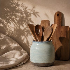 May include: A light blue ceramic utensil crock with a dark grey base and a stamped logo, filled with various wooden kitchen utensils including a slotted spoon, a solid spoon, and flat spatulas, positioned next to a wooden cutting board.