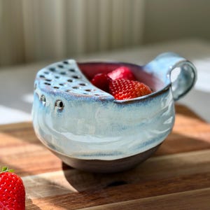 May include: A handmade ceramic berry bowl with a light blue exterior and a dark brown base. The bowl has a handle and a section with multiple holes. Fresh red strawberries are inside the bowl, which sits on a wooden cutting board.