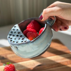 May include: A ceramic berry bowl with a light blue glazed finish and a dark brown base. The bowl features a built-in strainer section with multiple circular drainage holes on one side and a small handle. Several fresh red strawberries are inside.