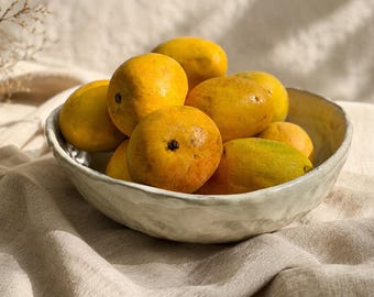 May include: A round, off-white ceramic bowl with a textured, uneven rim filled with a cluster of ripe, yellow-orange mangoes. The fruit has smooth, rounded skins with subtle variations in color, ranging from bright yellow to hints of green.