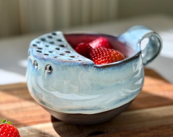 May include: A ceramic berry bowl with a light blue and white glazed exterior and a dark brown base. The bowl features a built-in colander section with circular drainage holes and a side handle. Fresh red strawberries are inside the bowl.