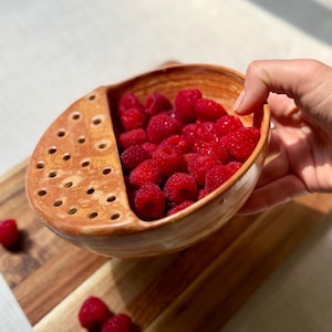 May include: A handmade, ceramic berry bowl filled with bright red raspberries. The bowl is a warm, earthy brown with a perforated section for draining. The bowl is on a wooden chopping board.