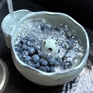 May include: A light blue ceramic colander bowl with an irregular rim and a central sculpted figure resembling a small creature, filled with fresh blueberries being rinsed under a stream of water from a faucet, creating bubbles and splashing.