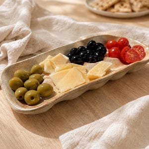 May include: An elongated, off-white ceramic serving tray with a wavy, organic edge. The tray is divided into three sections containing green olives, sliced hard cheese, black olives, and halved cherry tomatoes, arranged in a row.