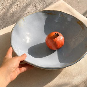 May include: A large, shallow, light blue ceramic bowl with an irregular, organic rim and subtle gold-toned accents along the edge. A single, round, deep red pomegranate with a brown stem sits in the center of the bowl, casting a distinct shadow.
