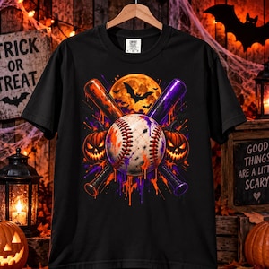 May include: A black short-sleeve t-shirt featuring a graphic print of a baseball centered between two crossed baseball bats. The design includes two glowing jack-o'-lanterns, a full moon, bats, and orange and purple paint drip effects.
