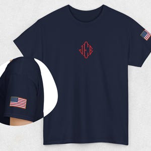 May include: Navy blue t-shirt featuring a small red embroidered design and an American flag patch on the sleeve. The shirt has a crew neck design.