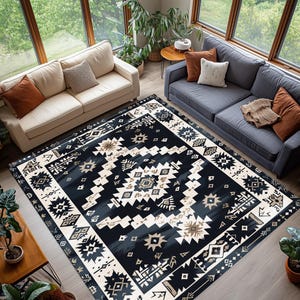 May include: A large, rectangular area rug with a geometric pattern in shades of navy, cream, and tan. The rug features a central design with repeating shapes and a border with similar motifs. The rug is placed in a living room setting with sofas and plants.