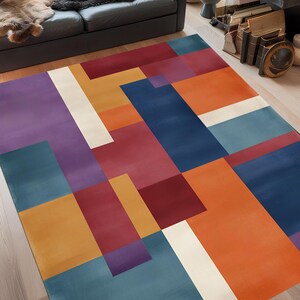 May include: A colorful geometric area rug with a modern design. The rug features a pattern of overlapping rectangles in shades of blue, orange, red, purple, and white. The rug is placed on a hardwood floor in a living room setting.