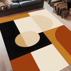 May include: A modern area rug with a geometric design. The rug features a combination of black, cream, tan, and rust-orange shapes, including squares, rectangles, and semi-circles. The rug is placed on a hardwood floor.
