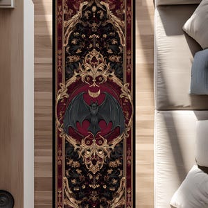 Pode incluir: Um tapete retangular com uma borda preta, apresentando um design central de um morcego com asas abertas dentro de um círculo vermelho. O tapete tem um fundo escuro com padrões dourados e pretos ornamentados.