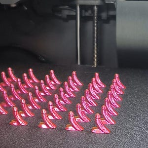 Puede incluir: Una base de impresora 3D con múltiples objetos pequeños, rosados y curvos. Los objetos están dispuestos en filas sobre una superficie oscura. El mecanismo de la impresora es visible en el fondo.