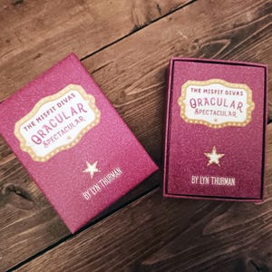 Puede incluir: Dos cajas rosas brillantes con el texto "The Misfit Divas Oracular Spectacular" y una estrella. Las cajas est&aacute;n sobre una superficie de madera. El nombre del autor, Lyn Thurman, est&aacute; impreso en la parte inferior de cada caja.