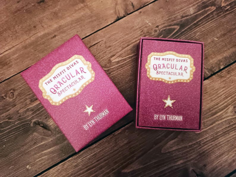Puede incluir: Dos cajas rosas brillantes con el texto "The Misfit Divas Oracular Spectacular" y una estrella. Las cajas est&aacute;n sobre una superficie de madera. El nombre del autor, Lyn Thurman, est&aacute; impreso en la parte inferior de cada caja.