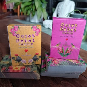 May include: Two pocket-size card decks, one oracle and one tarot. The oracle deck box is yellow with pink floral details and the words "Quiet Rebel Oracle." The tarot deck box is pink with "Quiet Rebel Tarot." Both are on floral card decks.