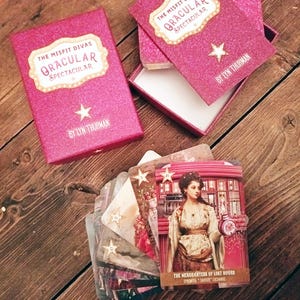 Puede incluir: Un juego de cartas de or&aacute;culo con purpurina rosa titulado "The Misfit Divas Oracular Spectacular" de Lyn Thurman. El juego incluye una baraja de cartas con ilustraciones de estilo vintage y una caja a juego.