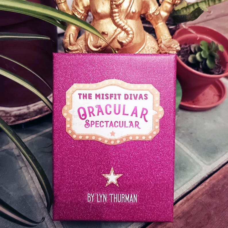 Puede incluir: Una caja rosa brillante con el texto "THE MISFIT DIVAS ORACULAR SPECTACULAR" en un dise&ntilde;o estilo marquesina. Debajo del texto hay una estrella blanca. La parte inferior de la caja dice "BY LYN THURMAN".