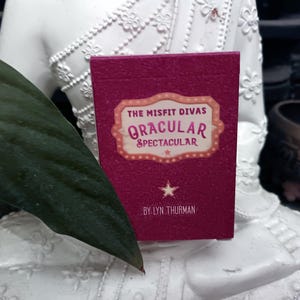 May include: A magenta-coloured box of "The Misfit Divas Oracular Spectacular" cards. The box features a cream-coloured label with the title and a star graphic. The author's name, Lyn Thurman, is printed at the bottom.