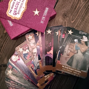 Puede incluir: Una baraja de cartas de or&aacute;culo titulada "The Misfit Divas Dracula Spectacular" con una caja burdeos brillante. Las cartas presentan ilustraciones con acentos de estrella. El texto "The Black Market Ballerina" es visible en una carta.