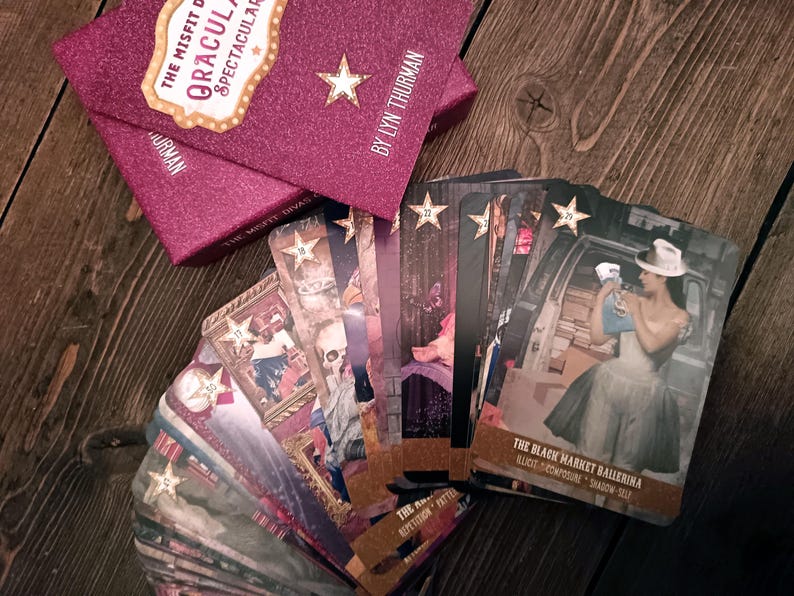 Puede incluir: Una baraja de cartas de or&aacute;culo titulada "The Misfit Divas Dracula Spectacular" con una caja burdeos brillante. Las cartas presentan ilustraciones con acentos de estrella. El texto "The Black Market Ballerina" es visible en una carta.