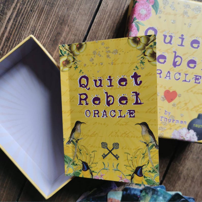 May include: A yellow box with the words "Quiet Rebel Oracle" printed in purple on the front. The box is decorated with birds, flowers, and keys. The box is open and the inside is white.