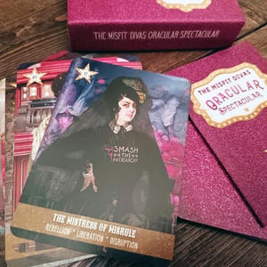 May include: An oracle card deck and box set. The cards display illustrations and text, including "THE MISTRESS OF MISRULE" and "SMASH THE PATRIARCHY". The box is pink with the text "THE MISFIT DIVAS ORACULAR SPECTACULAR". The cards and box are on a wooden surface.