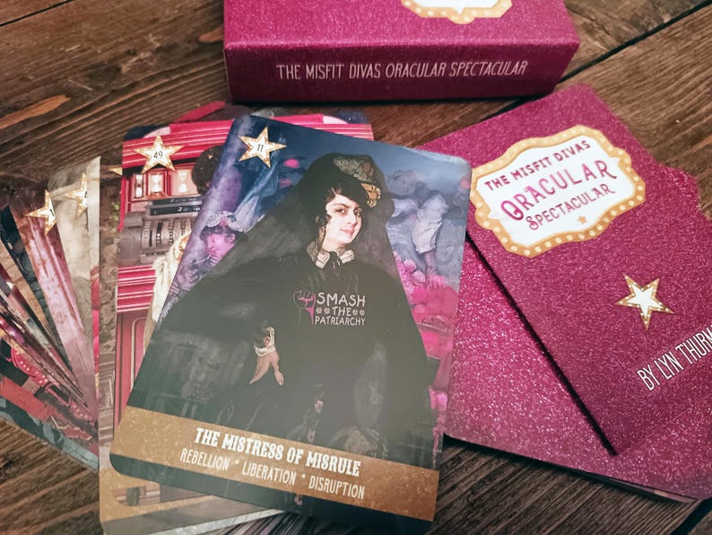 Puede incluir: Un mazo de cartas de or&aacute;culo y una caja rosa con el texto "THE MISFIT DIVAS ORACULAR SPECTACULAR". Las cartas presentan ilustraciones y texto, incluyendo "THE MISTRESS OF MISRULE" y "SMASH THE PATRIARCHY".