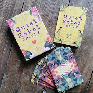 May include: A yellow box with the text "Quiet Rebel Oracle" and a heart symbol. The box is open and shows a deck of cards with colorful floral and bird illustrations. The cards are titled "Quiet Rebel Oracle" by Lyn Thurman.
