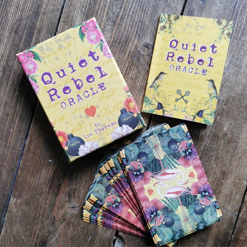 May include: A yellow box with the text "Quiet Rebel Oracle" and a heart symbol. The box is open and shows a deck of cards with colorful floral and bird illustrations. The cards are titled "Quiet Rebel Oracle" by Lyn Thurman.