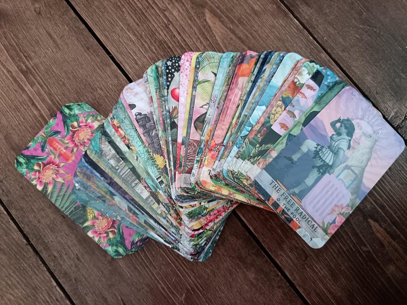 May include: A deck of tarot cards fanned out on a wooden surface. The cards feature colorful illustrations with floral, botanical, and figurative designs. One card displays the text "THE FREE RADICAL O THE FOOL."