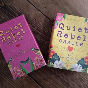 Puede incluir: Dos barajas de cartas, un tarot y un oráculo. La baraja "Quiet Rebel Tarot" es magenta con letras doradas, un corazón rojo e ilustraciones florales y de pájaros. La baraja "Quiet Rebel Oracle" es amarilla con letras moradas, un corazón rojo e ilustraciones florales.