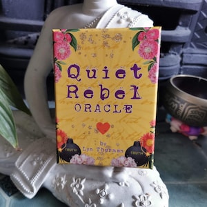 May include: A yellow and gold oracle deck box with the text "Quiet Rebel Oracle" in purple lettering. The box has a red heart and the text "by Lyn Thurman" in black lettering. The box is decorated with floral designs and black bombs with the word "Truth" on them.