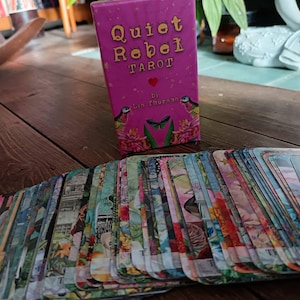 May include: A deck of tarot cards with a pink box that reads "Quiet Rebel Tarot." The cards are spread out, displaying colorful illustrations. The box features a heart, birds, and a butterfly design.