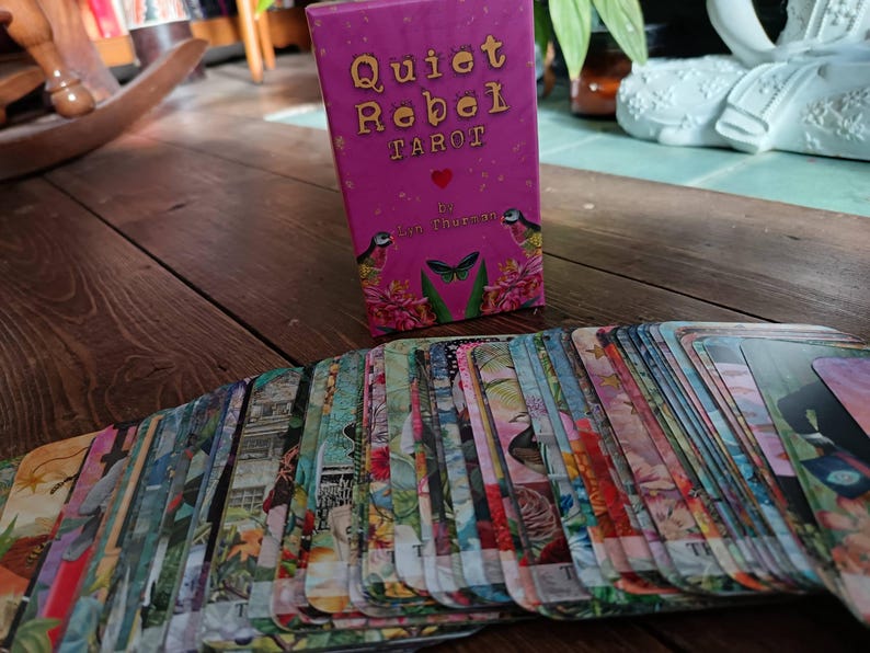 May include: A deck of tarot cards with a pink box that reads "Quiet Rebel Tarot." The cards are spread out, displaying colorful illustrations. The box features a heart, birds, and a butterfly design.