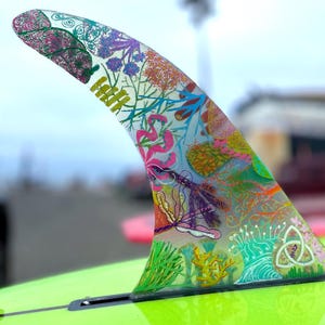 Op de afbeelding: Een kleurrijke surfplankvin met een bloemen- en abstract ontwerp. De vin is transparant en heeft een verscheidenheid aan kleuren, waaronder roze, groen, geel en blauw.
