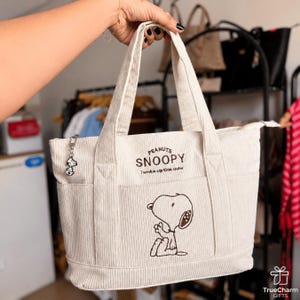 May include: A cream-colored corduroy tote bag featuring an embroidered Snoopy character on the front. The bag includes the text PEANUTS SNOOPY and Twinkie up this outer, with a small metal Snoopy charm attached to the zipper pull of the main compartment.
