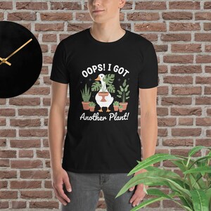 Peut inclure: T-shirt noir avec le texte "Oops! I Got Another Plant!" et un dessin d'une oie tenant un pot de plante, entourée de plantes en pot. Le t-shirt est porté par une personne.