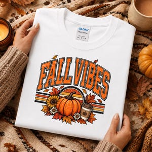Peut inclure: Un t-shirt blanc avec un motif imprimé portant le texte FALL VIBES en lettres orange à contour noir. Le design comprend une citrouille orange centrale, une tasse à café, des tournesols, des marguerites et des feuilles d'automne sur un fond rayé.