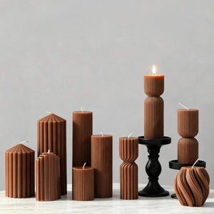 Peut inclure: Diverses bougies piliers marron de différentes formes. Certaines sont cylindriques, d'autres ont des motifs géométriques, et l'une est une spirale torsadée. Une bougie est allumée, et certaines sont sur des bougeoirs noirs. Les bougies sont sur une surface blanche.