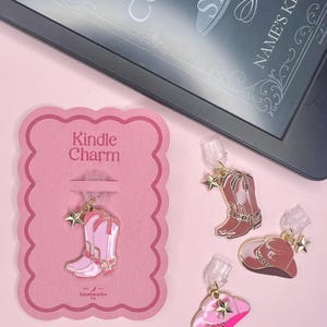 May include: A selection of Kindle charms featuring cowboy boot and hat designs in shades of pink and brown. One charm is attached to a pink card with the text "Kindle Charm". A Kindle e-reader is visible in the background.