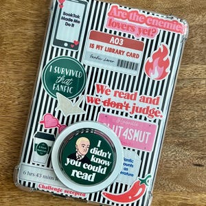 May include: Clear phone case with a black and white striped background, covered in stickers. Stickers include text like "BookTok Made Me Do It," "I Survived that Fanfic," and "Are the enemies lovers yet?" A pop socket with a portrait and the text "I didn't know you could read" is also visible.