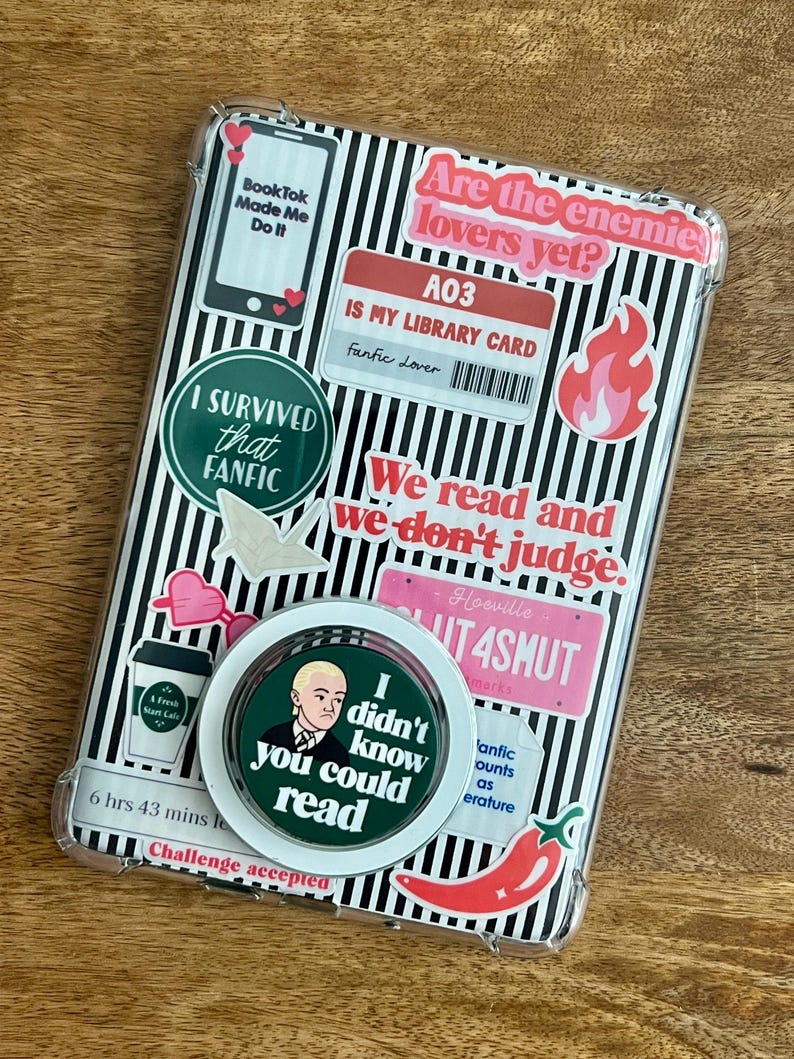 May include: Clear phone case with a black and white striped background, covered in stickers. Stickers include text like "BookTok Made Me Do It," "I Survived that Fanfic," and "Are the enemies lovers yet?" A pop socket with a portrait and the text "I didn't know you could read" is also visible.
