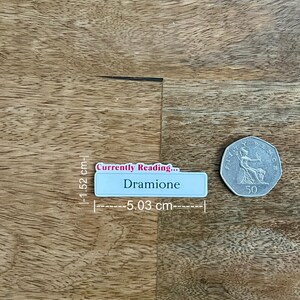 May include: A light blue rectangular name tag, 2 inches long, with the text "Currently Reading... Dramione". A silver 50 pence coin is next to the name tag. The objects are on a wooden surface.