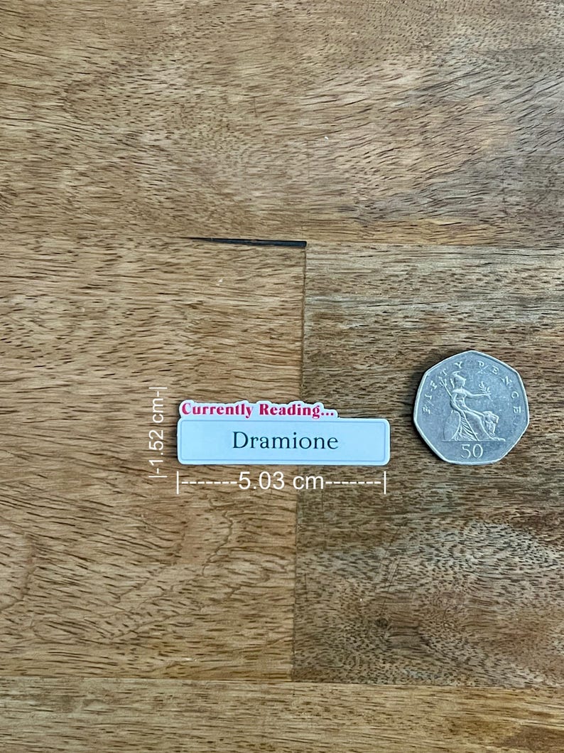 May include: A light blue rectangular name tag, 2 inches long, with the text "Currently Reading... Dramione". A silver 50 pence coin is next to the name tag. The objects are on a wooden surface.