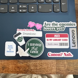 May include: A collection of stickers on a laptop, including a pink heart-shaped glasses sticker, a sticker that says "Are the enemies lovers yet?", and a sticker that says "I survived that fanfic".