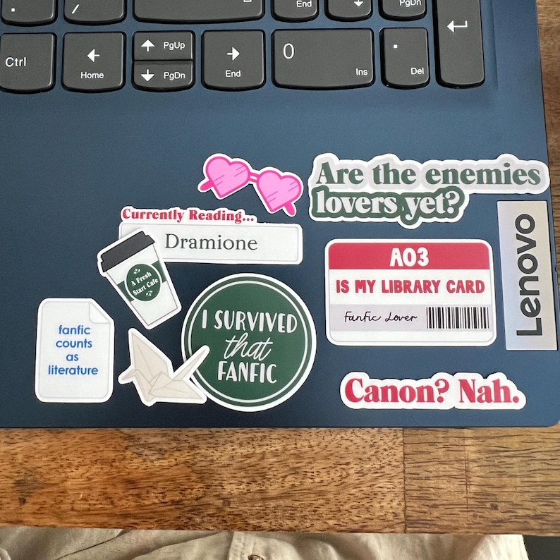 May include: A collection of stickers on a laptop, including a pink heart-shaped glasses sticker, a sticker that says "Are the enemies lovers yet?", and a sticker that says "I survived that fanfic".