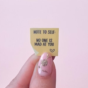 Può includere: Un piccolo post-it giallo con il testo "NOTE TO SELF: NO ONE IS MAD AT YOU" e un simbolo a forma di cuore. Il biglietto è tenuto in mano con smalto rosa e un disegno floreale dorato.