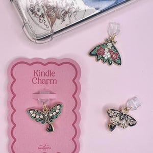 May include: Three Kindle charms with butterfly designs. One charm is attached to a pink card that reads "Kindle Charm". The charms have gold-colored metal accents and are decorated with black, white, green, and red enamel.