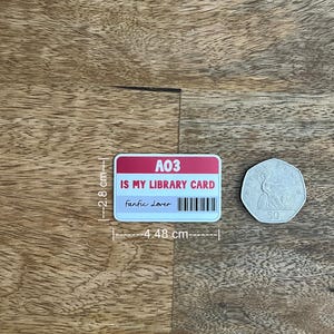May include: A rectangular sticker with the text "A03 IS MY LIBRARY CARD Fanfic Lover" and a barcode. The sticker is 4.48 cm wide and 2.8 cm tall. A 50 pence coin is next to the sticker.