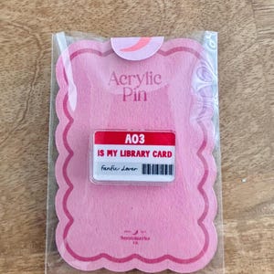 Puede incluir: Un pin acrílico rosa en una funda de plástico transparente. El pin tiene forma de tarjeta de biblioteca con el texto "A03 IS MY LIBRARY CARD fanfic lover" en blanco y negro. El embalaje tiene un borde festoneado.